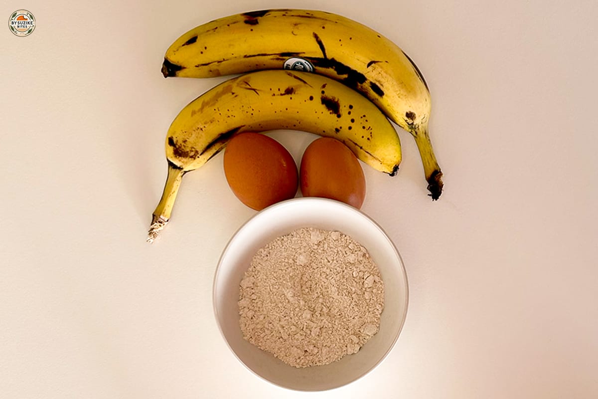 Oat Banana Cake 10 Two ripe bananas, two eggs, and a bowl of oat flour on a white background, ready for a healthy oat and banana cake recipe.